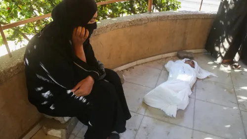 امرأة ترتدي الحجاب تجلس على حافة رصيف، تعبر عن الحزن بينما يستلقي طفل مغطى بغطاء أبيض على الأرض، في مشهد يبرز معاناة المجاعة في غزة.