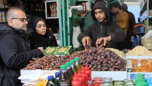مشهد لثلاثة أشخاص يتسوقون في سوق غزة، حيث يظهرون أمام أكوام من التمر وزجاجات زيت، مع بائع يقوم بتحضير الطلبات.
