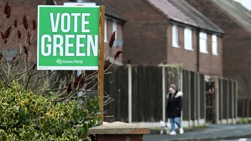 لافتة انتخابية لحزب الخضر مكتوب عليها "صوتوا للخضر"، مع مشاة في الخلفية يتجولون في حي سكني في مانشستر.