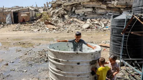 طفل فلسطيني يستحم في برميل ماء وسط أنقاض مبانٍ مدمرة في قطاع غزة، يعكس تداعيات الحرب المستمرة والدمار المدني في المنطقة.