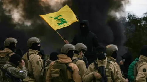 شخص يرتدي زي عسكري يحمل علم حزب الله الأصفر، محاطًا بجنود آخرين في موقع مظلم، مما يعكس التوترات السياسية في لبنان.