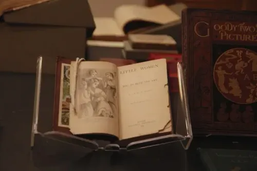 كتاب "نساء صغيرات" مفتوح على طاولة في متحف بوسطن أثينيوم، محاط بكتب تاريخية أخرى، يعكس إرث القراءة والثقافة.