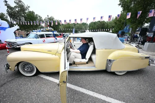 سيارة كلاسيكية صفراء مكشوفة متوقفة في حدث عام، مع شخص يجلس داخلها، وخلفها سيارات أخرى وزينة بأعلام أمريكية.