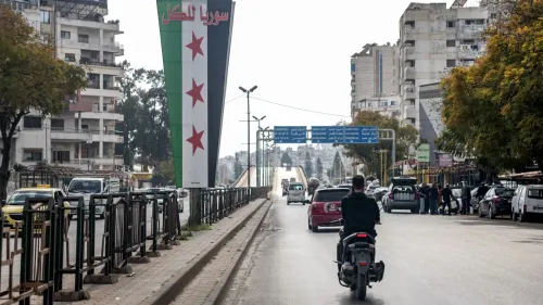 افتتاحية لمدينة سورية تظهر لافتة كبيرة تحمل نجوم حمراء على خلفية العلم الوطني، مع مشهد حضري يتضمن سيارات وأشخاص في الشارع.