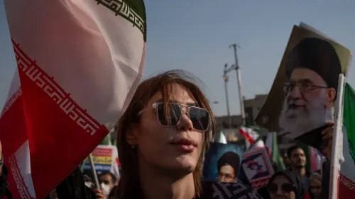 امرأة ترتدي نظارات شمسية وتحمل علم إيران، بينما يتواجد متظاهرون آخرون في الخلفية يحملون صورًا للمرشد الأعلى علي خامنئي.