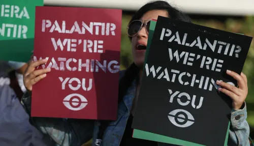 امرأة تحمل لافتة مكتوب عليها "Palantir We're Watching You" خلال احتجاج، تعبيراً عن القلق من انتهاكات حقوق الإنسان.