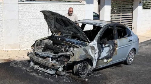 سيارة محترقة مع غطاء محرك مفتوح، بينما يقف رجل بجانبها، تعكس آثار العنف والاعتداءات في المنطقة.