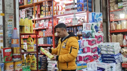 رجل يرتدي سترة صفراء يتفقد زجاجة زيت في متجر مليء بالمنتجات الغذائية والسلع الاستهلاكية، مع تكدس الأرفف بالسلع.