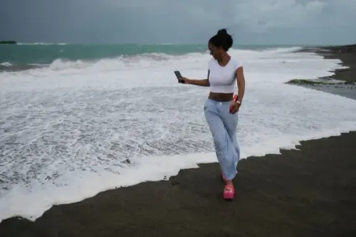 امرأة تسير على الشاطئ بالقرب من أمواج عاتية، تحمل هاتفًا محمولًا، في ظل أجواء غائمة نتيجة اقتراب الإعصار ميليسا.