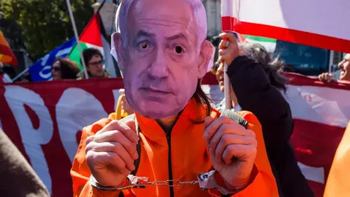 شخص يرتدي زيًا برتقاليًا ويمسك بأصفاد، يحمل قناعًا يمثل رئيس الوزراء الإسرائيلي بنيامين نتنياهو، وسط مظاهرة.
