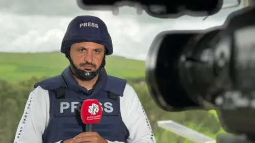 صحفي يرتدي سترة وخوذة واقية تحمل علامة "PRESS"، يتحدث أمام كاميرا في جنوب لبنان، مع خلفية طبيعية.