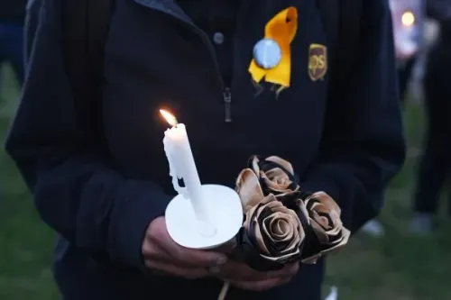 شخص يحمل شمعة مضاءة وزهورًا مصنوعة من الورق، ويرتدي سترة سوداء مع شارة صفراء، في تجمع تأبيني لضحايا حادث تحطم طائرة UPS.