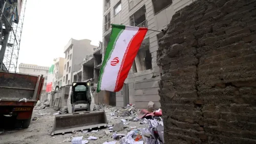 علم إيران يرفرف في موقع دمار، مع وجود حطام المباني وآثار الدمار حوله، مما يعكس تأثير الغزو المستمر على البلاد.