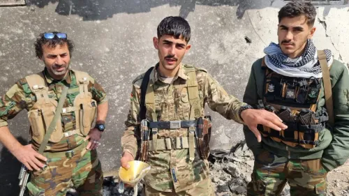 ثلاثة مقاتلين كرديين يقفون أمام حائط متضرر، يرتدون زيًا عسكريًا ويظهرون أسلحة، في سياق التوترات الحالية في إيران.