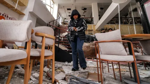 امرأة ترتدي سترة داكنة تتجول بين حطام مطعم دمرته الغارات الجوية في طهران، مع كراسي مكسورة وأنقاض حولها.