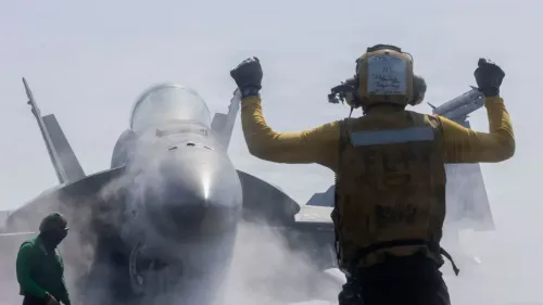 صورة لعسكري يوجه طائرة مقاتلة من طراز F/A-18 في قاعدة بحرية، مع تصاعد الدخان حول الطائرة، مما يعكس التوترات العسكرية في المنطقة.