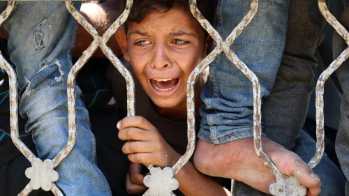 طفل فلسطيني يبكي ويصرخ بين قضبان حديدية، مع وجود أقدام أخرى بالقرب منه، مما يعكس معاناة المدنيين في غزة خلال الصراع.