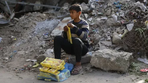 طفل فلسطيني يجلس على الرصيف وسط أنقاض وركام، ممسكًا بعلب طعام، مما يعكس معاناة المدنيين في غزة خلال التصعيد المستمر.