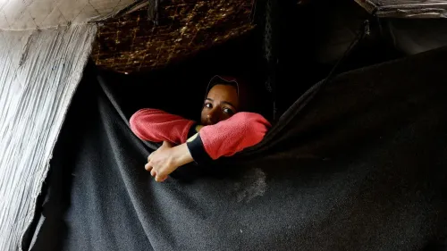 طفلة فلسطينية تنظر من خلف ستار في مخيم، تعكس معاناة الأطفال في ظل الأوضاع الإنسانية الصعبة في غزة.