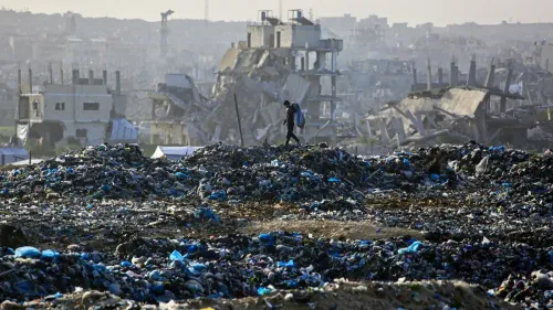 رجل يسير فوق كومة من النفايات في غزة، مع خلفية لمبانٍ مدمرة، تعكس الأوضاع الإنسانية الصعبة في المنطقة.
