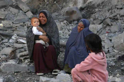 نساء وأطفال يجلسون على أنقاض مبنى مدمر في غزة، مع تعبيرات حزن واضحة على وجوههم، في ظل أزمة إنسانية متفاقمة.