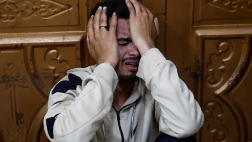 رجل يجلس على الأرض، يضع يديه على رأسه ويبدو عليه الحزن الشديد، في خلفية جدار خشبي. تعكس الصورة الألم الناتج عن الأحداث الأخيرة في غزة.