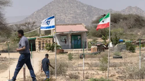 رجلان يسيران بالقرب من مبنى صغير يحمل علم إسرائيل وعلم أرض الصومال، في منطقة ذات تضاريس مرتفعة.