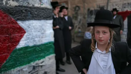 طفلة ترتدي ملابس تقليدية يهودية تقف أمام جدار مزين بألوان علم فلسطين، مما يعكس التوترات السياسية والاجتماعية في المنطقة.