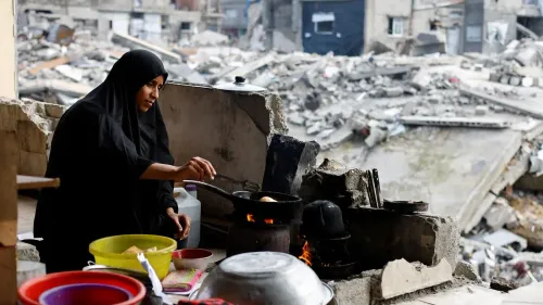 امرأة ترتدي الحجاب تطبخ في مطبخ بدائي وسط أنقاض مبانٍ مدمرة في غزة، تعكس معاناة السكان في ظل الظروف الصعبة.