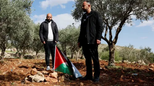 رجلان فلسطينيان يقفان بين أشجار الزيتون، أحدهما يحمل علم فلسطين، تعبيرًا عن المقاومة في ظل تصاعد العنف من المستوطنين.