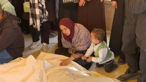 امرأة ترتدي حجابًا تجلس بجانب طفل صغير، وتظهر علامات الحزن على وجهها، بينما يحيط بهم أشخاص آخرون في موقع جنازة، مع وجود جثث مغطاة بأقمشة بيضاء.