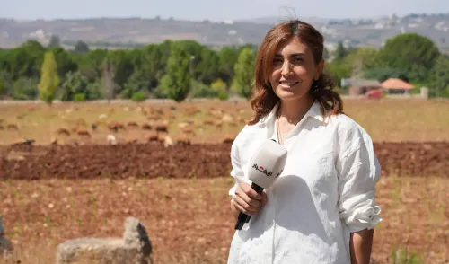 صحفية لبنانية تحمل ميكروفون، تبتسم في حقل زراعي، مع خلفية من الأشجار والتلال، تعكس مشهد الحياة اليومية في لبنان.