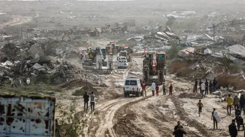 تظهر الصورة مشهدًا من منطقة مدمرة، حيث تتواجد آليات ثقيلة وسيارات إسعاف، مع أشخاص يتجولون في الأرجاء. تعكس الصورة آثار النزاع وتأثيره على المدنيين.