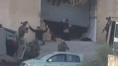 مواجهات في جنين تظهر فلسطينيين غير مسلحين يرفعان أيديهما، بينما تتواجد قوات الاحتلال الإسرائيلية في الخلفية.