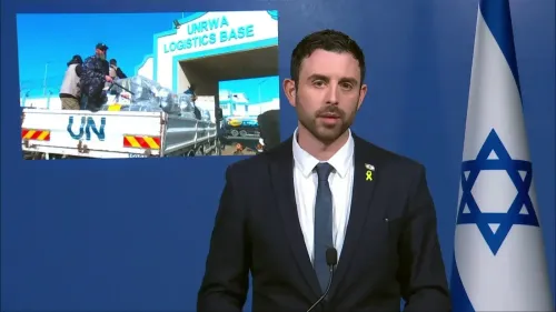 مستشار إسرائيلي يتحدث في مؤتمر صحفي، مع صورة خلفية تظهر قاعدة لوجستية تابعة للأونروا، تعكس جهود الدعاية الإسرائيلية.