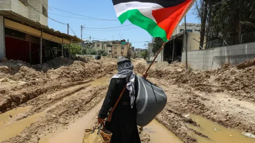 رجل يحمل علم فلسطين واقفًا في شارع مغمور بالمياه والطين، مما يعكس التحديات التي تواجه الفلسطينيين في الضفة الغربية.