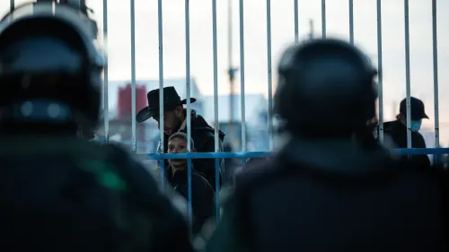 مشهد من مخيم الهول يظهر محتجزين يتطلعون عبر السياج، بينما يرتدي الجنود ملابس مكافحة الشغب. يعكس الوضع المتوتر في المخيم.