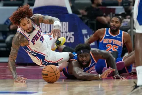 لاعب كرة سلة من فريق فيلادلفيا يسقط على الأرض أثناء المنافسة مع لاعب من نيويورك نيكس، مع كرة سلة بينهما.