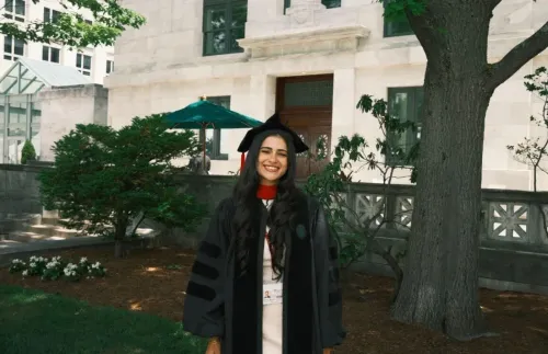 لين عزّ الدين، خريجة كلية الطب في Harvard، تبتسم بفخر مرتديةً زي التخرج، مع خلفية من الأشجار والمباني الأكاديمية.