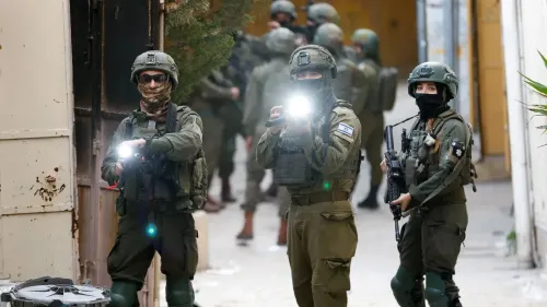 جنود إسرائيليون يرتدون زيًا عسكريًا كاملًا، يحملون أسلحة ومصابيح، أثناء قيامهم بدورية في منطقة حضرية، مع خلفية تضم مباني ومجموعة من الجنود الآخرين.