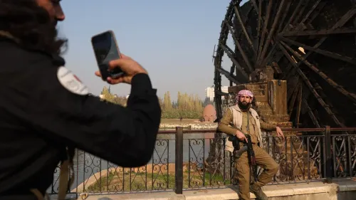 رجل مسلح يرتدي زيًا عسكريًا يقف بجوار عجلة مائية تاريخية في حماة، بينما يصور شخص آخر المشهد بهاتفه.