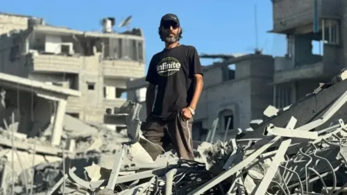 رجل يقف وسط أنقاض منزله المدمر في غزة، مع خلفية لمبانٍ مهدمة، يعكس آثار النزاع المستمر والألم الذي يعاني منه السكان.