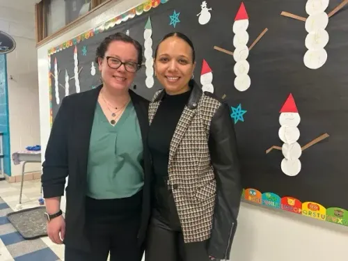 صورة لامرأتين مبتسمتين تقفان معًا في ممر مدرسة، خلفهما جدار مزين برموز شتوية، مما يعكس جهود تحسين التعليم في مرحلة الطفولة المبكرة.