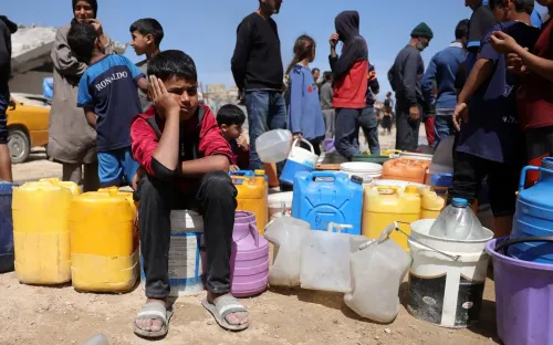 طفل يجلس على براميل بلاستيكية ملونة في غزة، بينما يتجمع الناس حوله لملء المياه، معبرين عن معاناتهم في ظل نقص الموارد الأساسية.