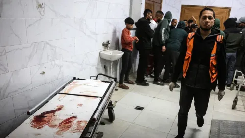 مشهد من مستشفى في غزة يظهر مسعفاً يرتدي سترة برتقالية، بينما يتجمع الناس في الخلفية حول سرير طبي مغطى بالدماء، مما يعكس تأثير النزاع على الصحة العامة.