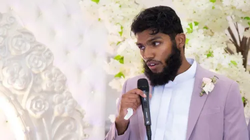 شاب يتحدث في مناسبة، يرتدي بدلة وردية مع قميص أبيض، ويظهر خلفه ديكور زهور بيضاء، يعكس أجواء احتفالية.