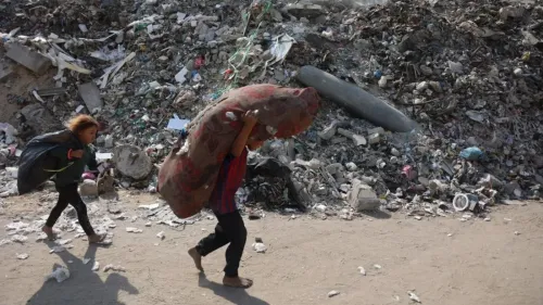 طفلان يحملان أمتعة فوق ظهورهم، يسيران بجانب كومة من النفايات في غزة، مما يعكس آثار النزاع المستمر والظروف الإنسانية الصعبة.