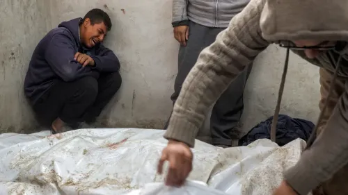 رجل شاب يجلس في زاوية غرفة، يبكي بحرقة، بينما يقوم شخص آخر بتغطية جثة ملفوفة في قماش أبيض، مما يعكس الفاجعة في غزة.