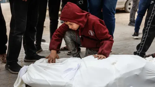 طفل يرتدي سترة حمراء ينحني فوق جثمان مغطى بغطاء أبيض، محاط بأشخاص في خان يونس، يعبر عن الحزن على فقدان أحد الأطفال بسبب البرد.