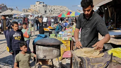شخص يعمل على إعداد الخبز في سوق مزدحم في غزة، حيث تظهر خلفه أنقاض المباني المدمرة وأشخاص يتسوقون في ظروف صعبة.